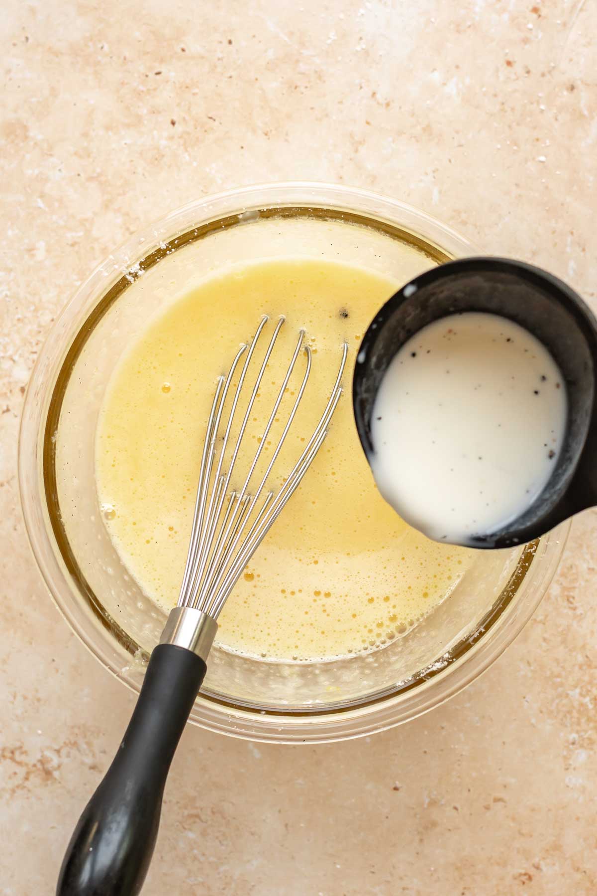 A ladle adds hot milk to eggs and sugar in a bowl.