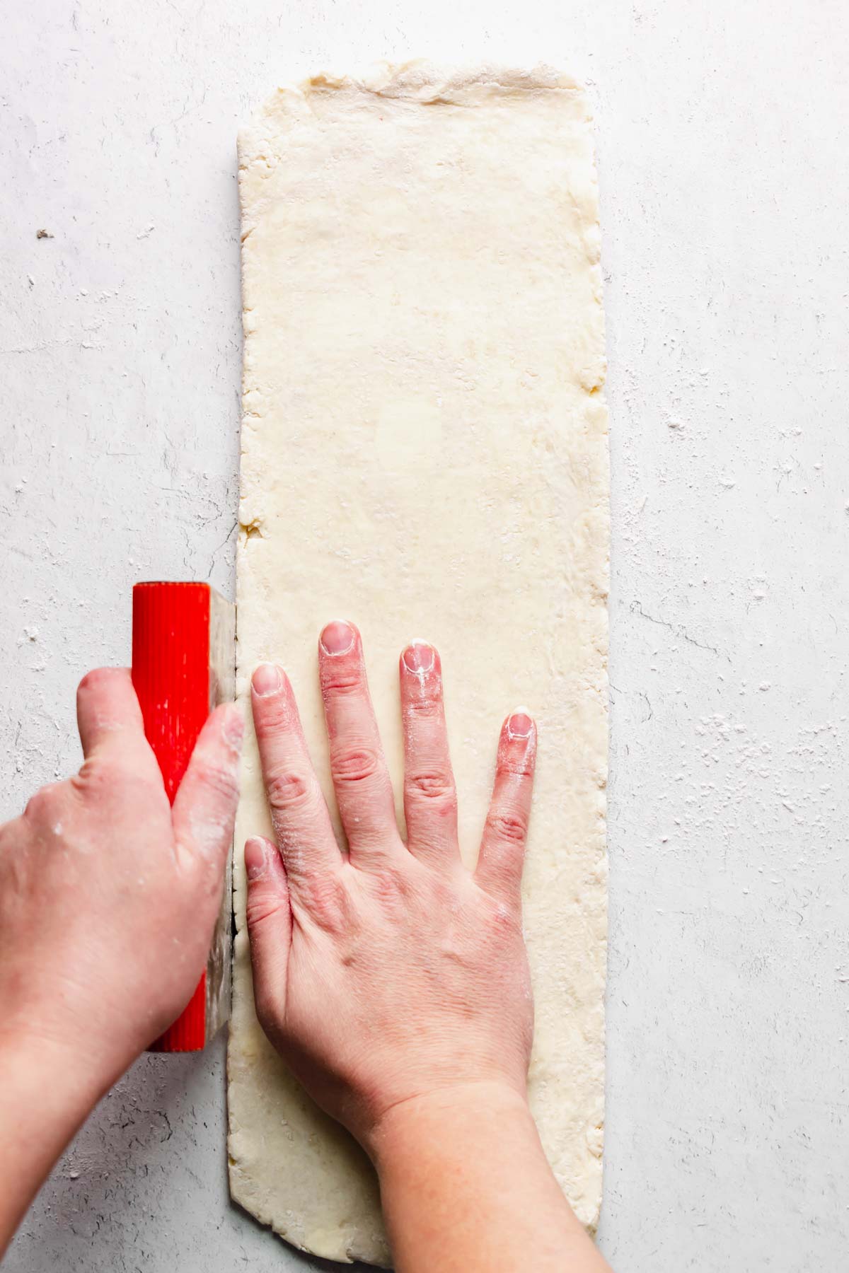 A long rectangle of dough with a hand and bench scraper cleaning up the sides.