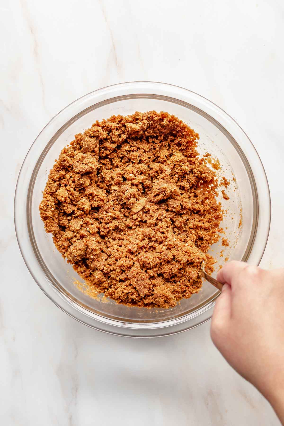 A hand mixes Biscoff cookie crumbs in a bowl.