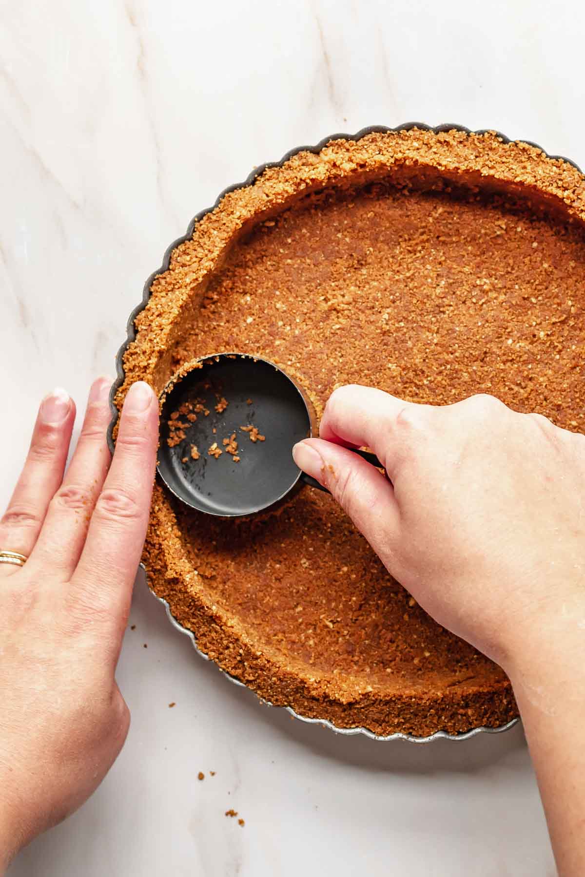 Hands use a measuring cup to press cookie crumbs into a tart pan.