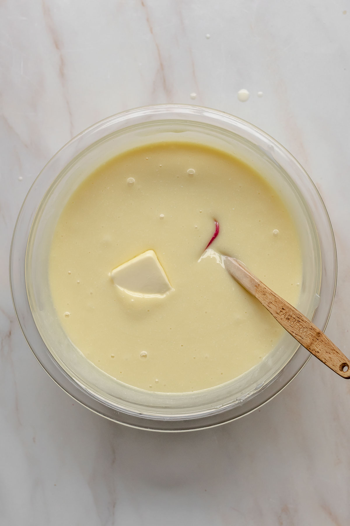 A pat of butter in the white chocolate ganache with a spatula resting in the bowl.