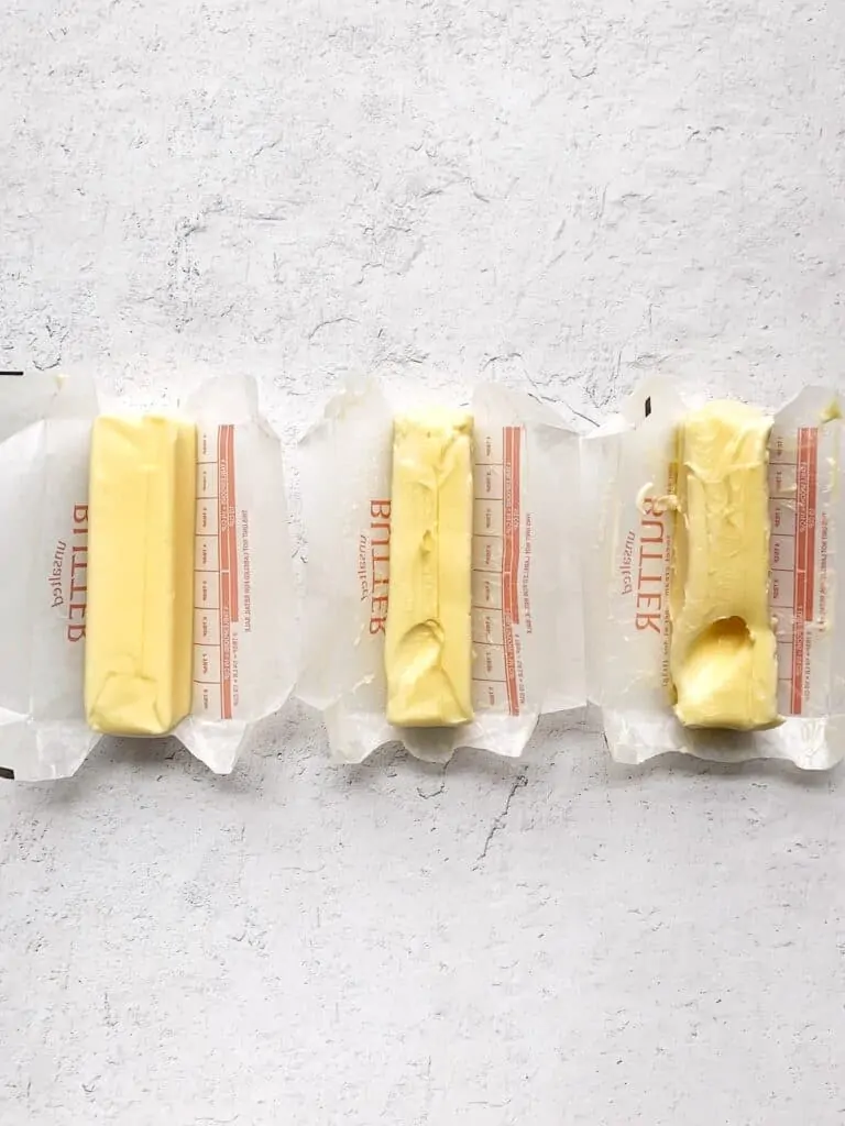 Three sticks of butter next to each other. One is cold, one at room temperature, one too soft. Each stick of butter has a finger indent.