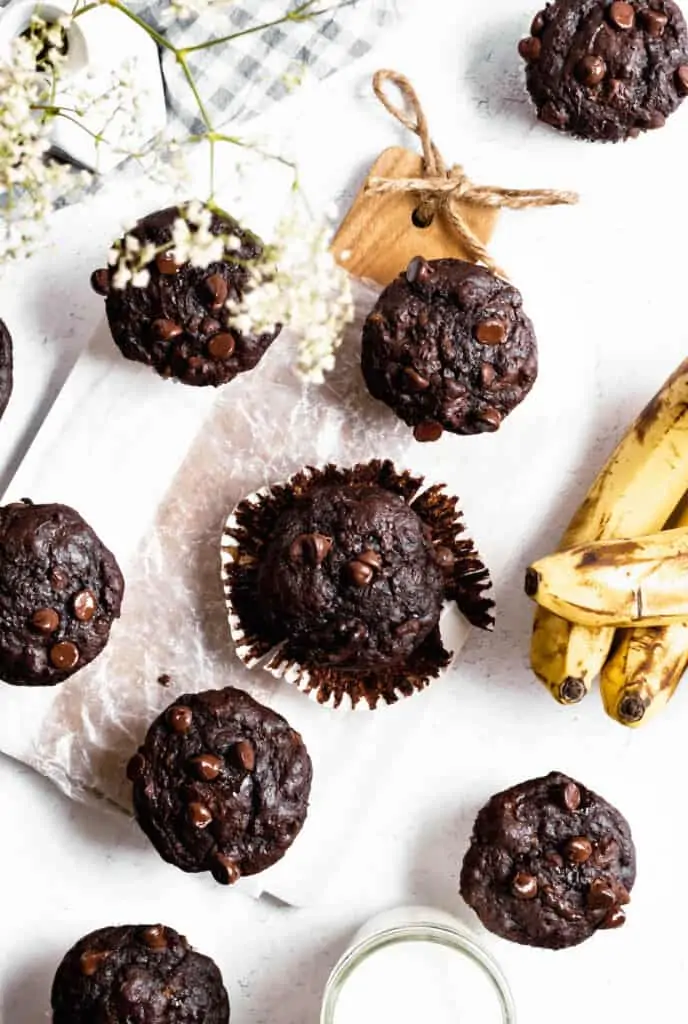 Overhead shot of numerous double chocolate banana muffins. The muffin in the center of the frame is unwrapped. Bananas and a glass of milk sit off on the side