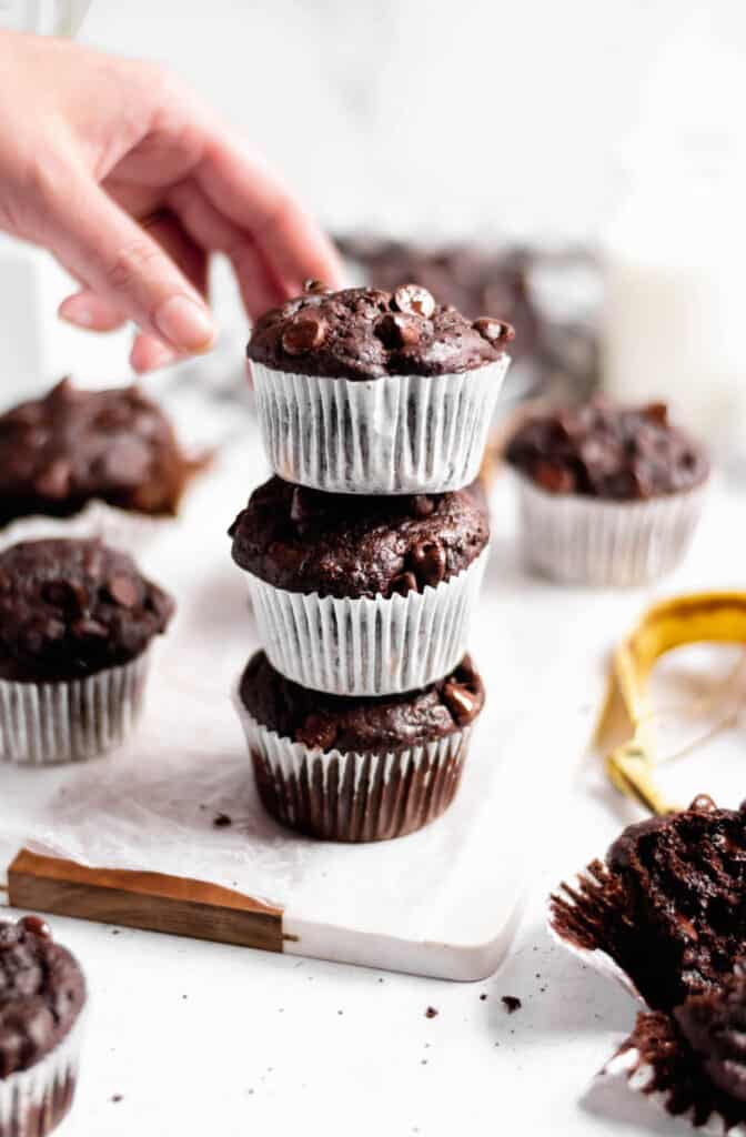 A stack of three double chocolate banana muffins. A hand reaches in to take the top muffin.