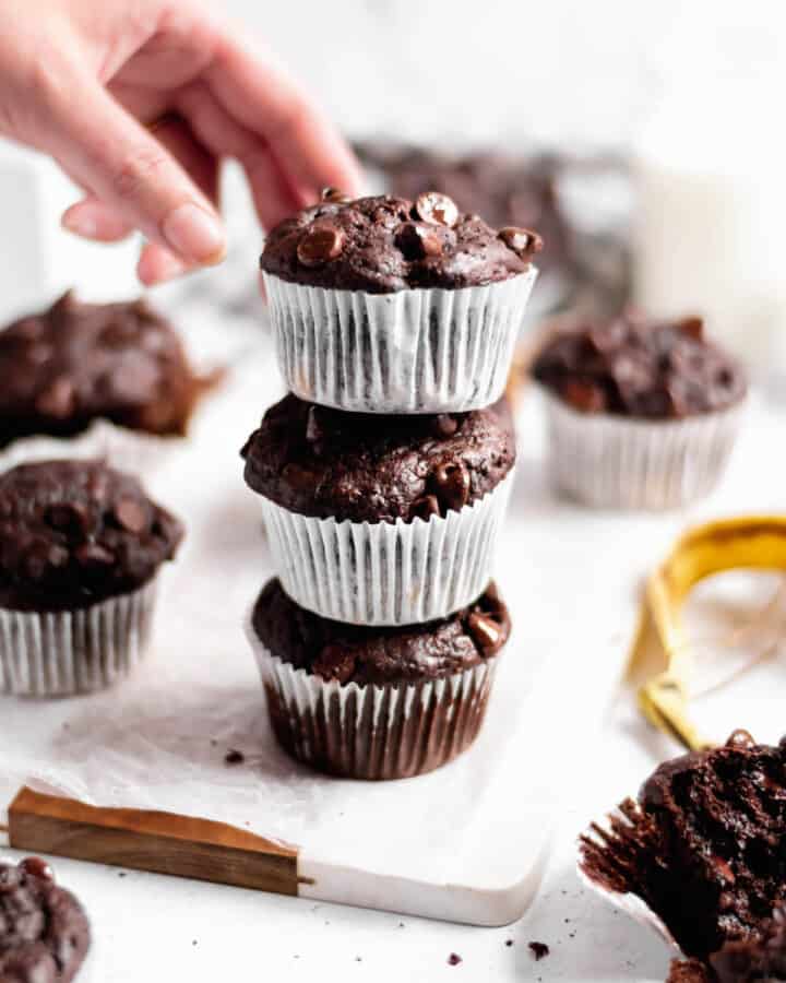 A stack of three double chocolate banana muffins. A had reaching in to take the top muffin.