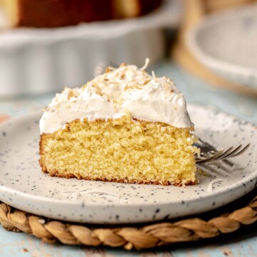 A slice of coconut almond cake on a plate with a fork next to it.