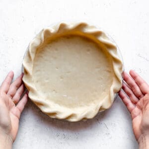 A crimped raw pie dough with two hands about to lift it up