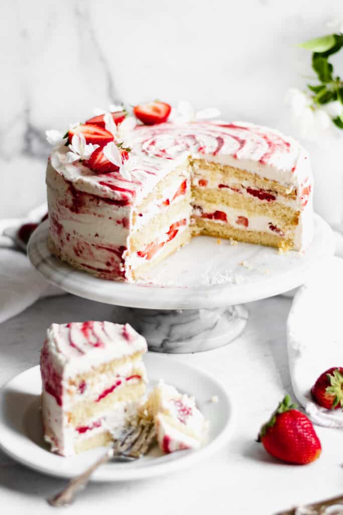 A slice of strawberry shortcake layer cake sits on a place with a bite removed. the full cake is placed on a cake stand with the insides exposed.