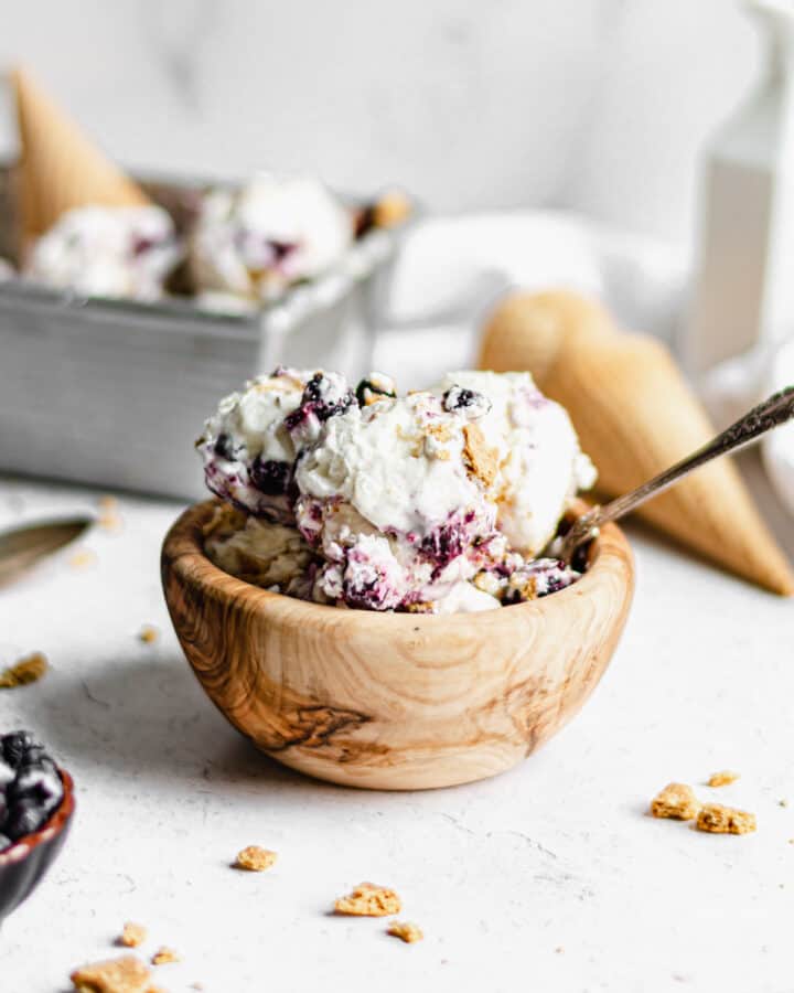 A wooden bowl filled with blueberry cheesecake no-churn ice cream. The container of ice cream sits int he background with ice cream cones.