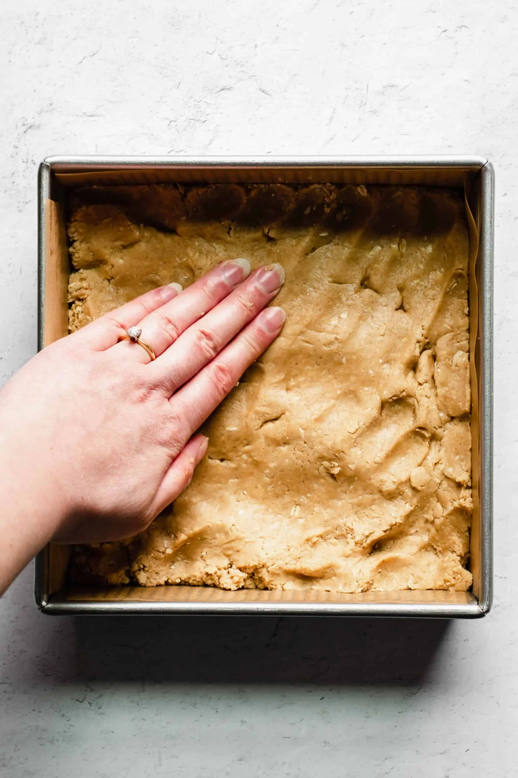 A hand presses peanut butter oat dough into a square pan.