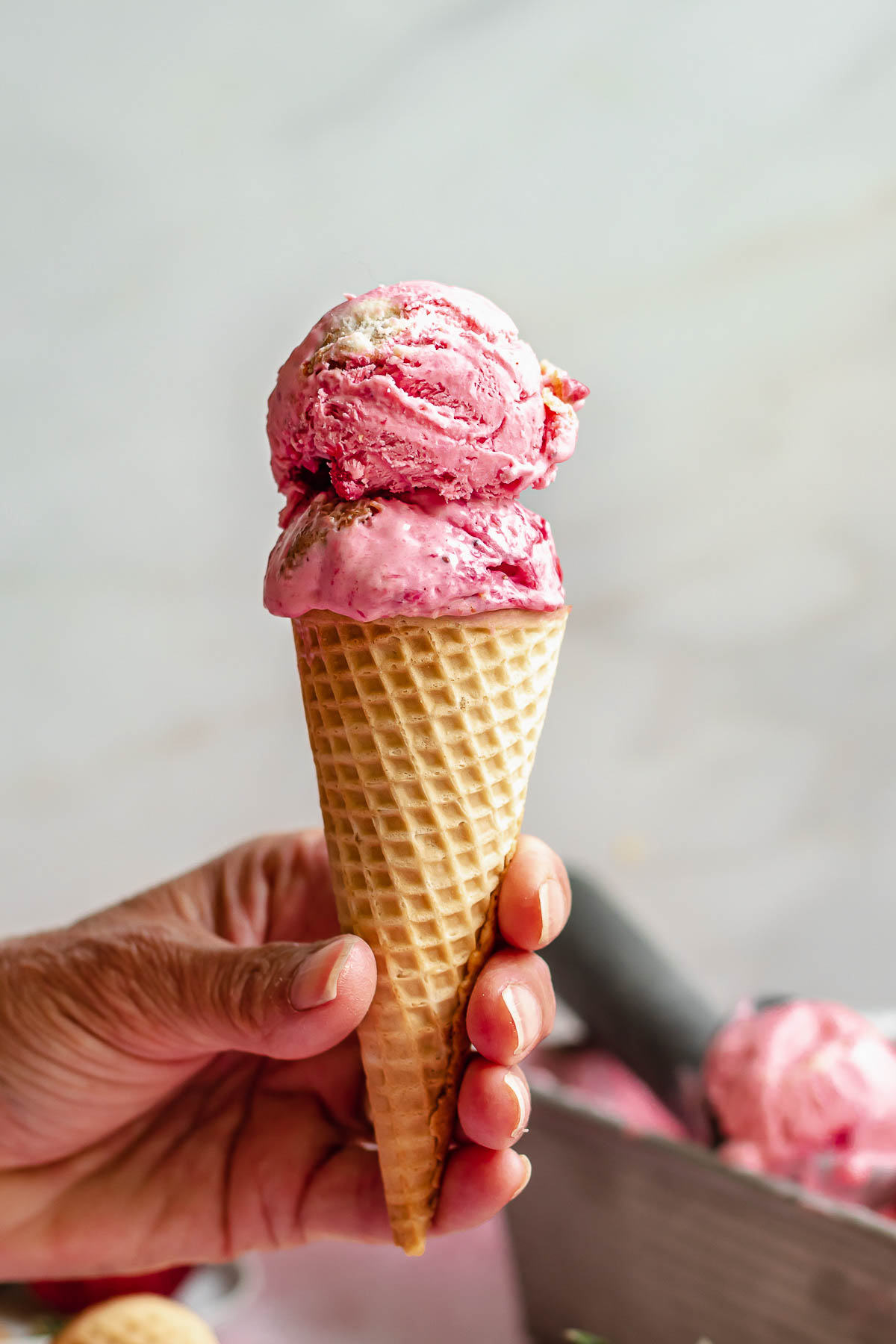 A hand holds an ice cream cone with two scoops of strawberry cheesecake ice cream in it.