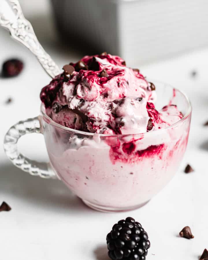 Straight on shot of blackberry chip ice cream in a glass coffee cup with a spoon in it. A fresh blackberry sits in front of the ice cream.