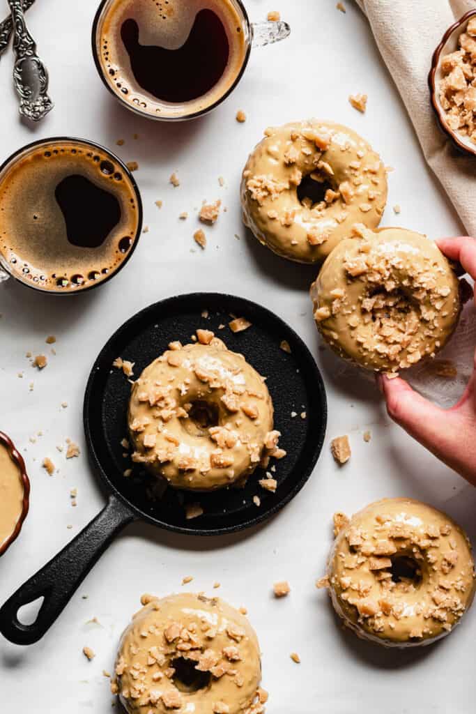 overhead shot of coffee toffee donuts with coffee cups. a hand reaches in to remove a donut