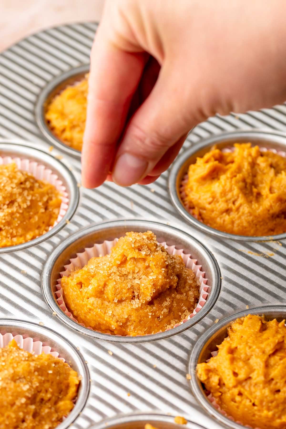 A hand adds coarse sugar to the tops of the muffin batter in the muffin tin.