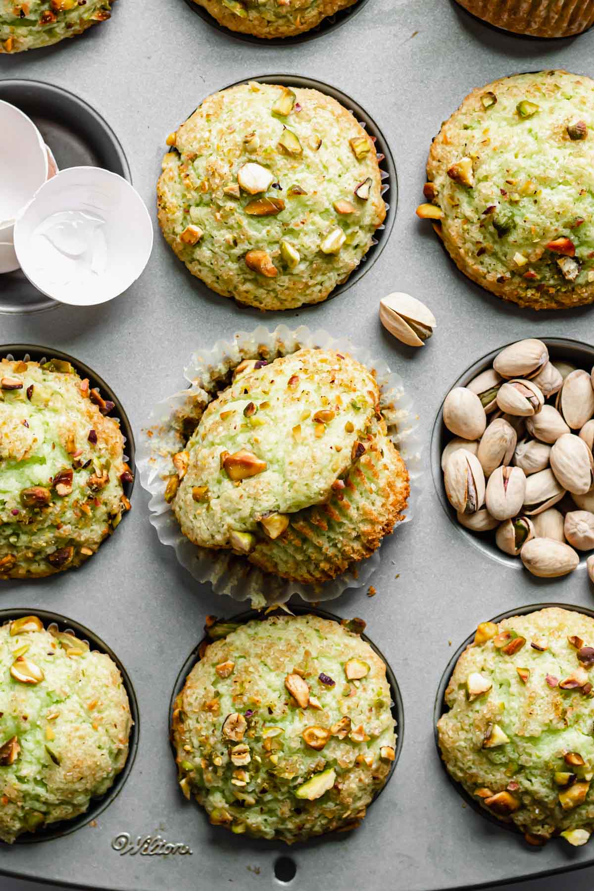 A muffin tin full of pistachio muffins. One muffin lays on its side and out of the liner next to a well of pistachios.