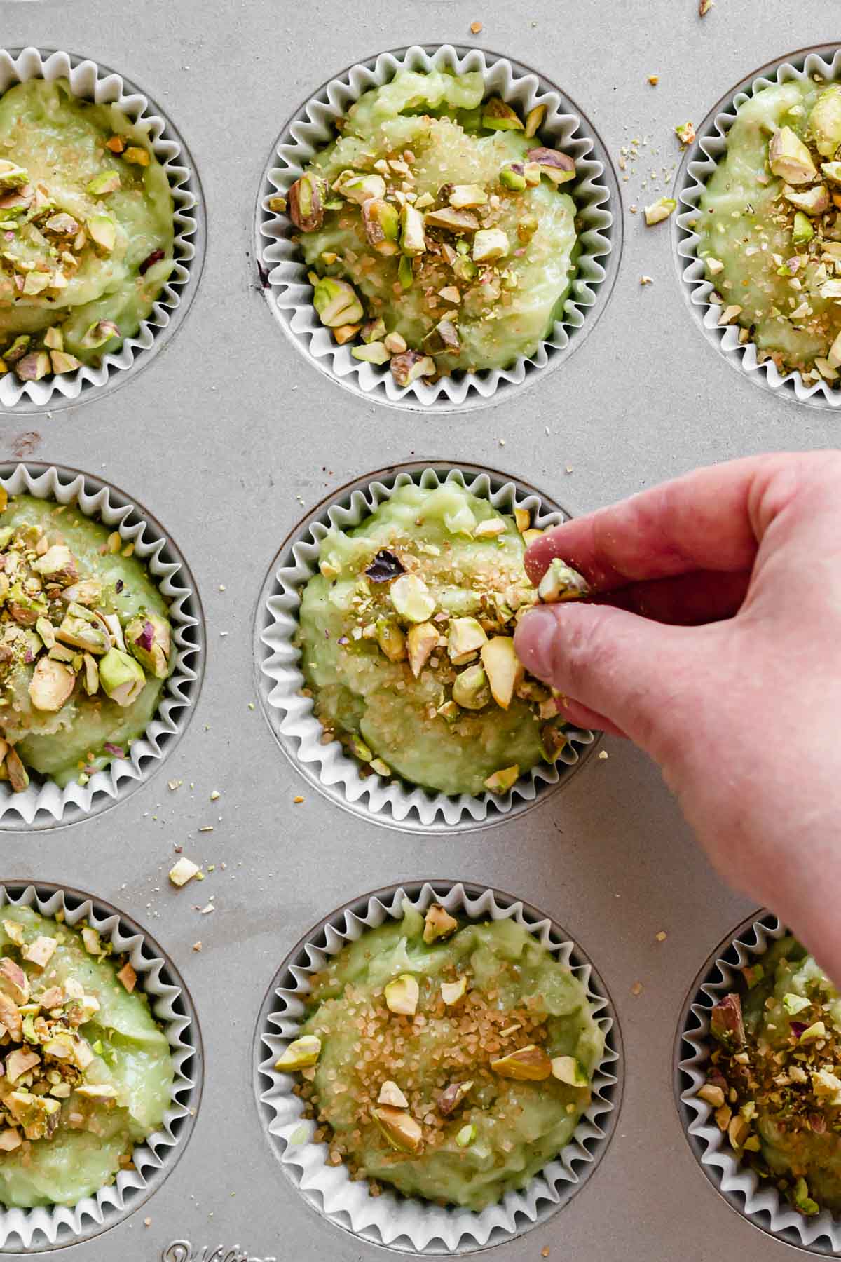 A hand adds chopped pistachios to the top of the muffin batter.