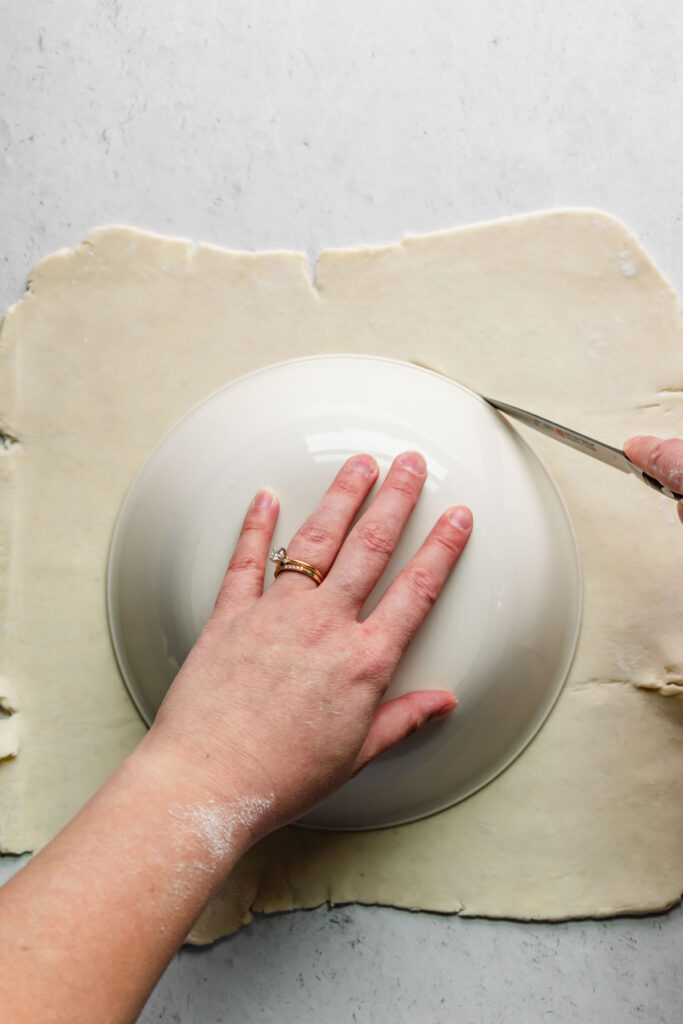 A bowl on top of rolled out rough puff pastry. A knife cuts alongside the bowl.