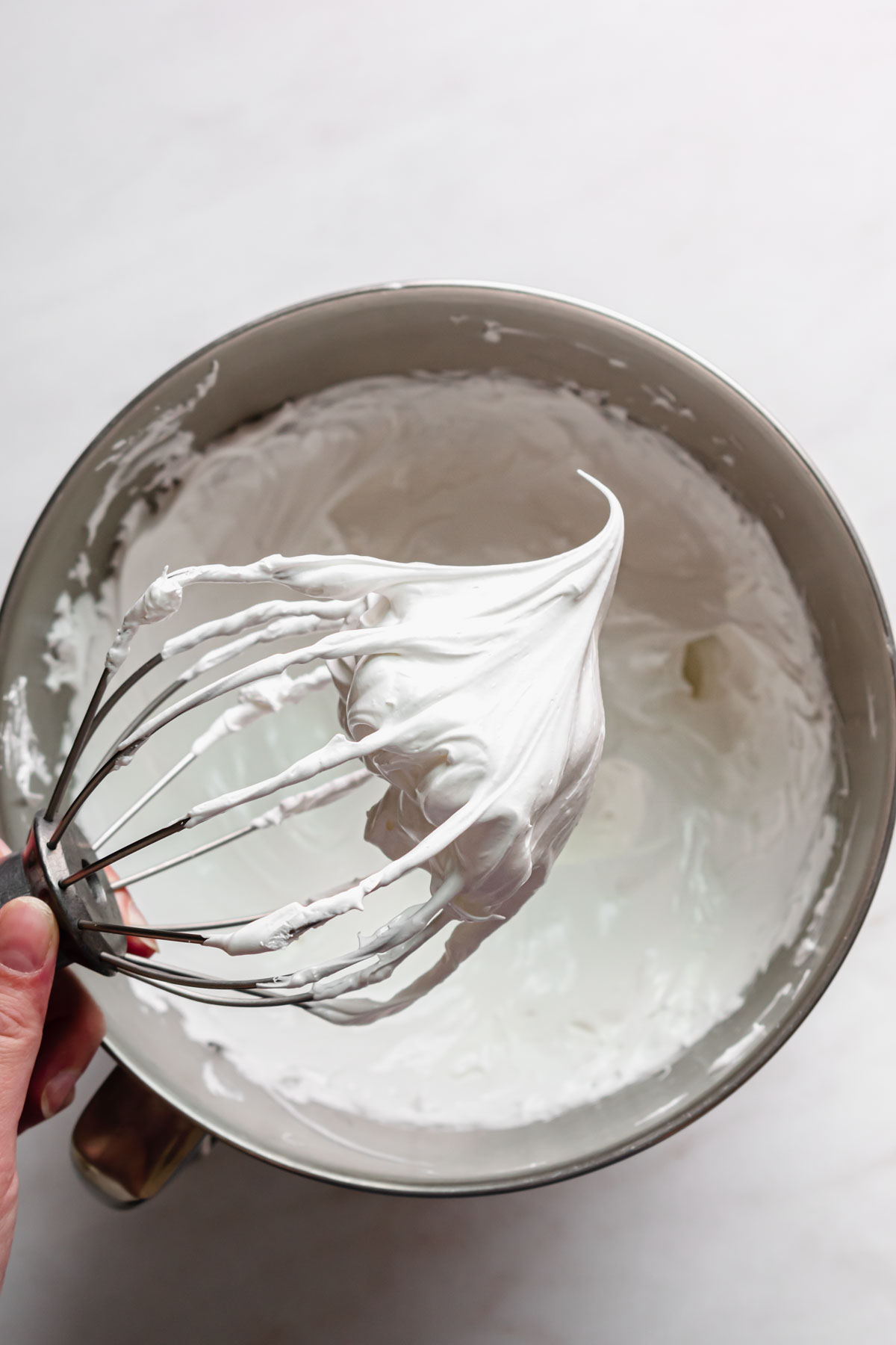 A whisk held out over bowl to show the stiff swiss meringue.