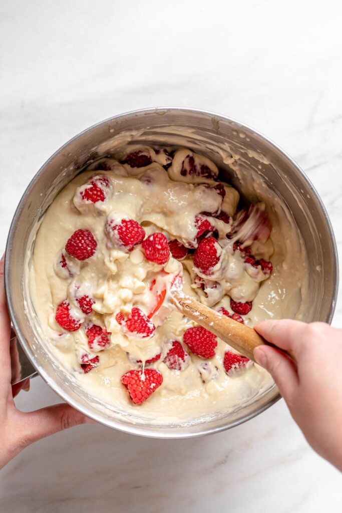 A hand folds the raspberries and white chocolate into the batter.