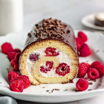 Swiss roll cake with a slice removed and raspberries scattered around the platter.