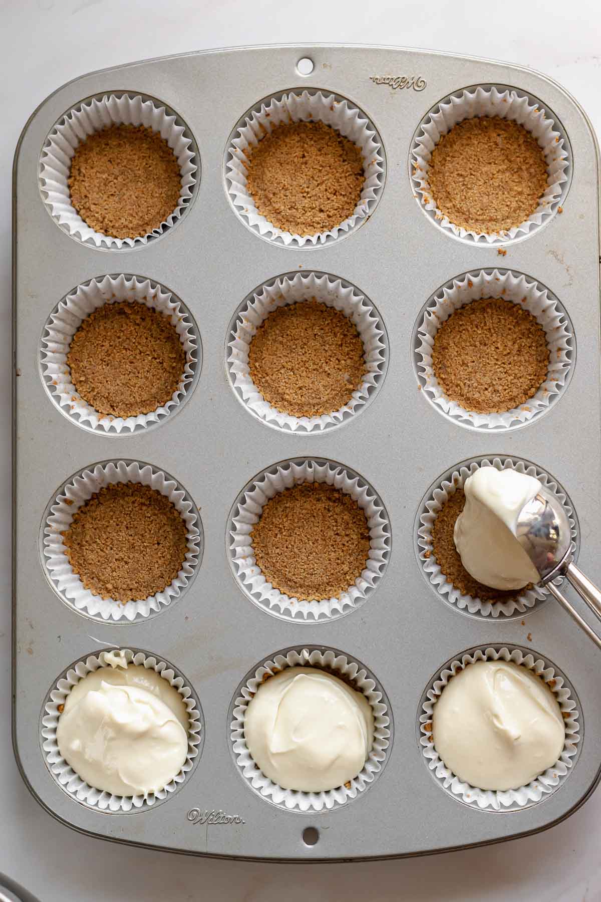 A cookie scoop adds cream cheese batter on top of the graham cracker crust.
