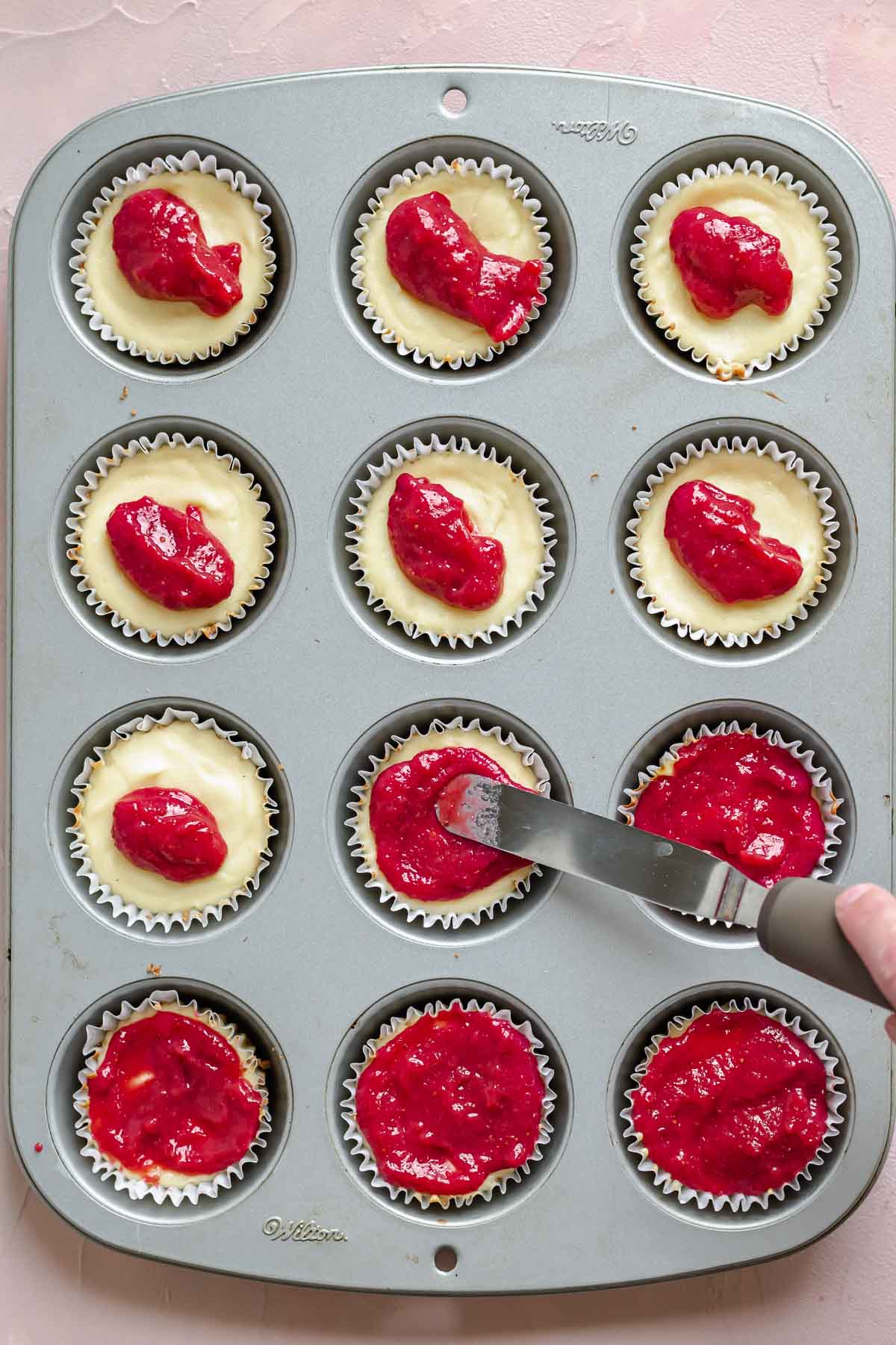 A rubber spatula spreads strawberry sauce onto baked mini cheesecakes.