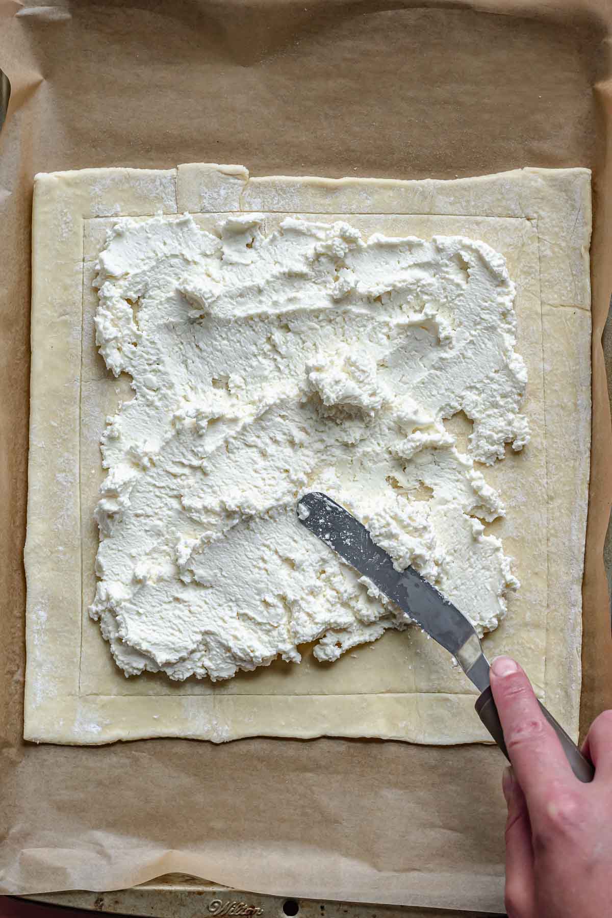 A spatula spreads cheese mixture onto puff pastry.