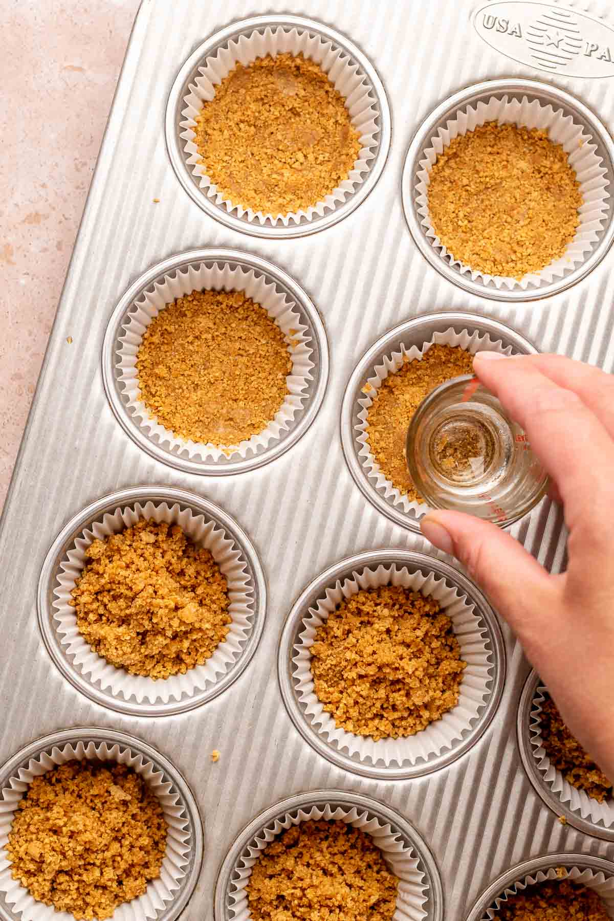 A hand uses a shot glass to press graham cracker crumbs into cupcake liners.