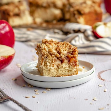 Slice of apple streusel coffee cake on a plate with the rest of the cake in the background.