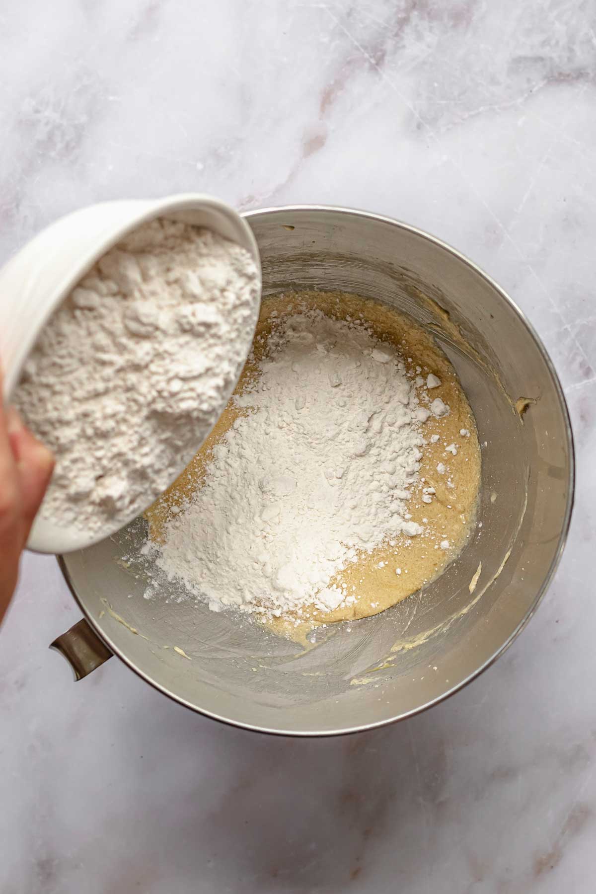 A hand pours a bowl of dry ingredients into wet ingredients.