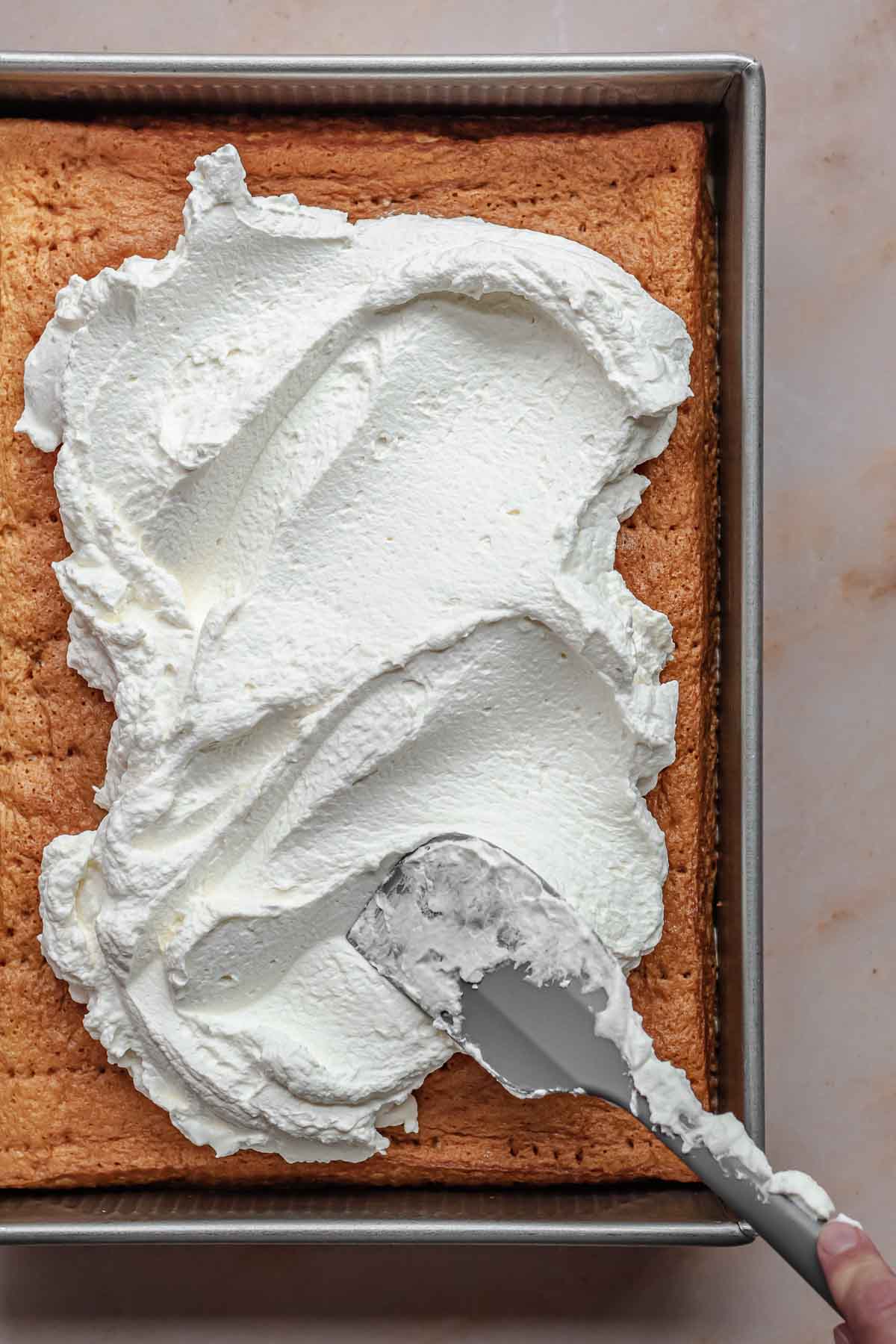 A spatula spreads fresh whipped cream onto the cake.