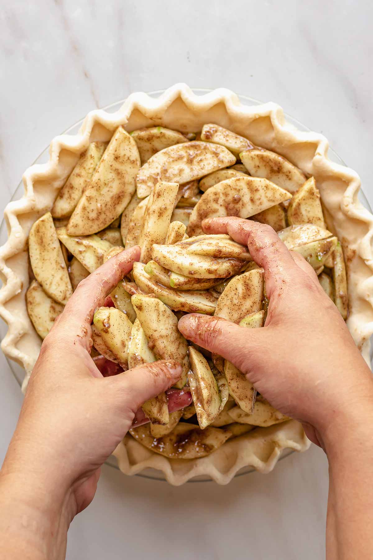 Hands adding spiced apple slices to a pie crust.