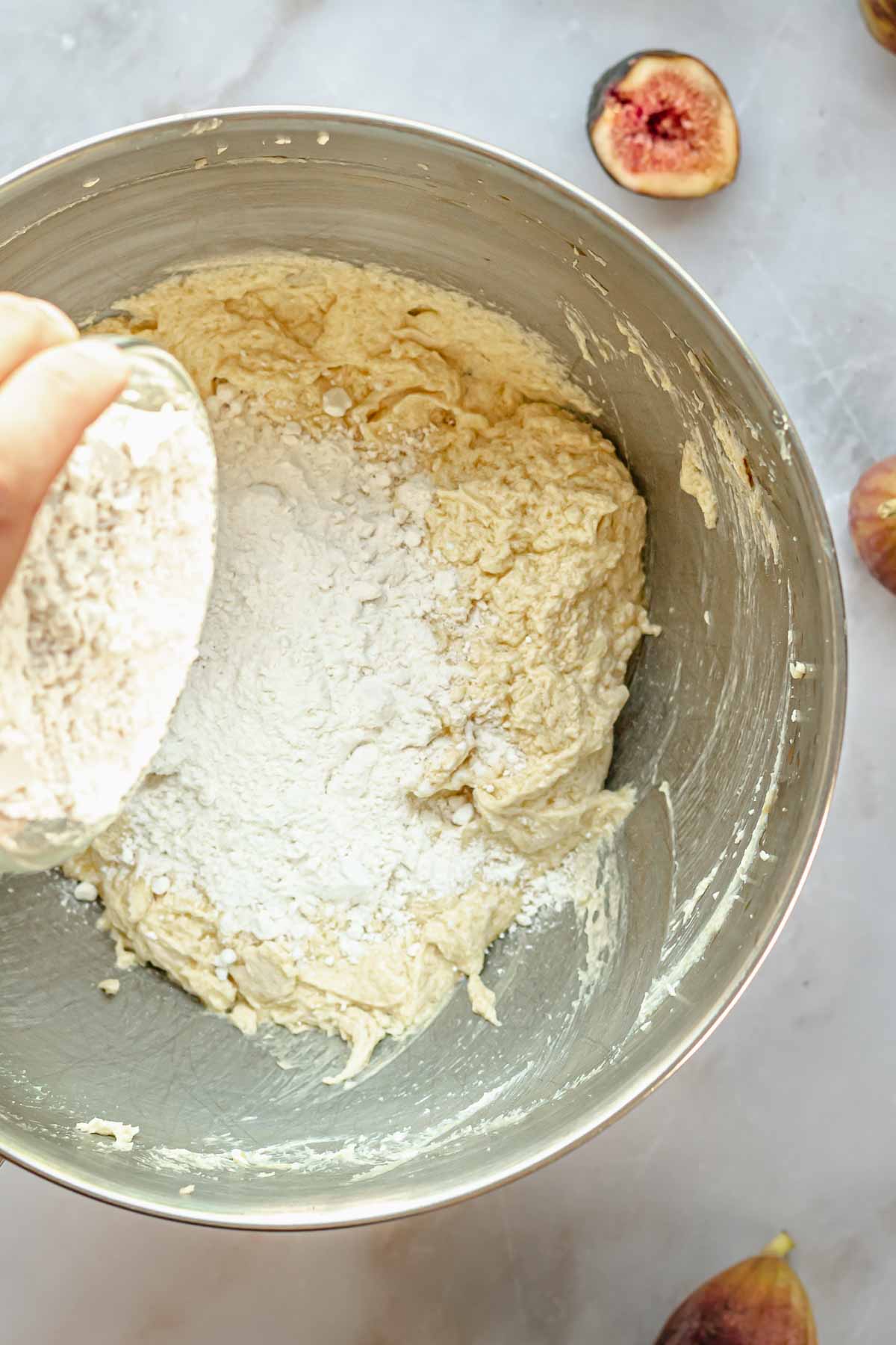 A hand pours flour into creamed butter and sugar.