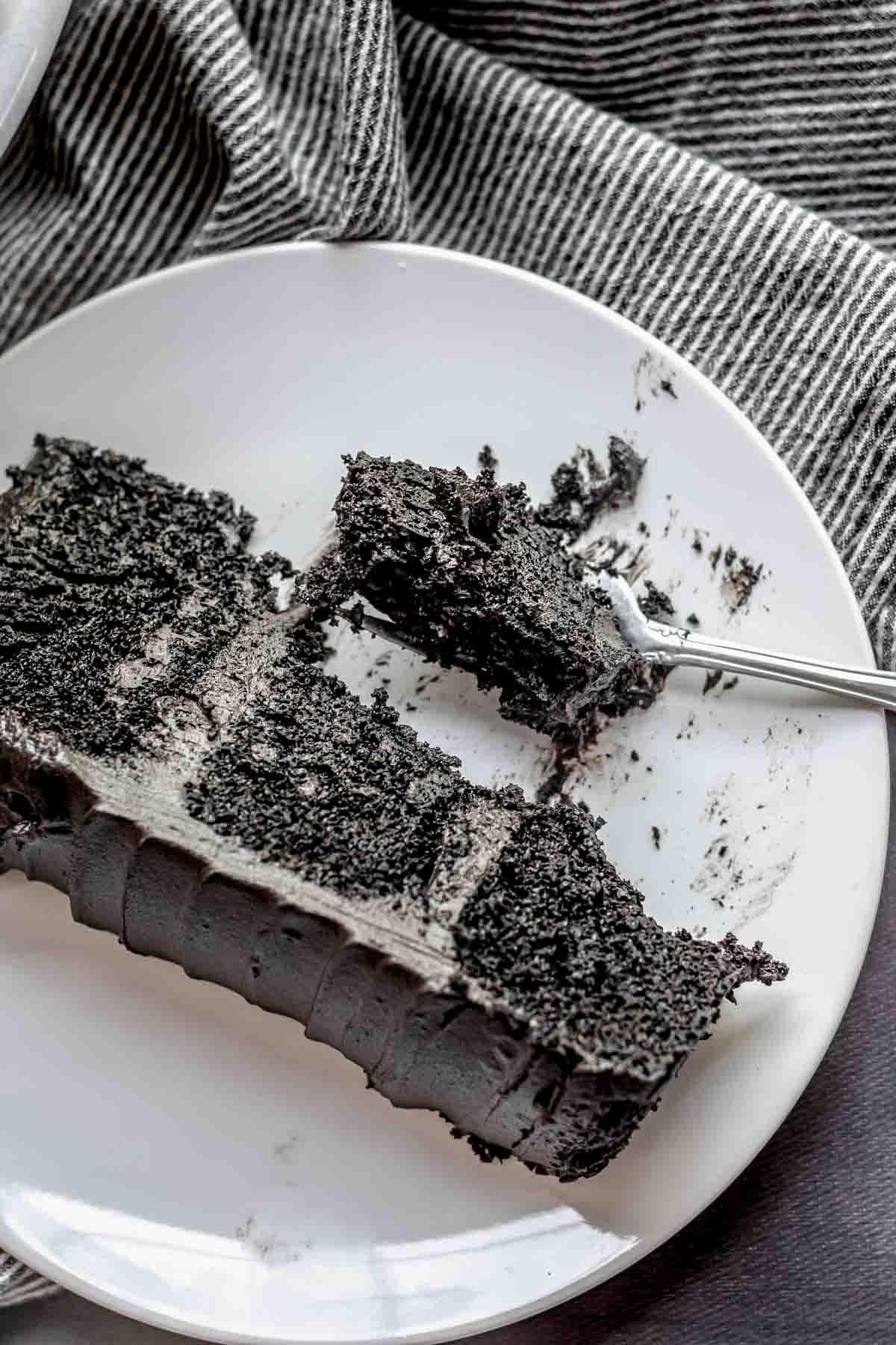 A slice of black cocoa cake on a plate. A fork has a piece of cake on it.