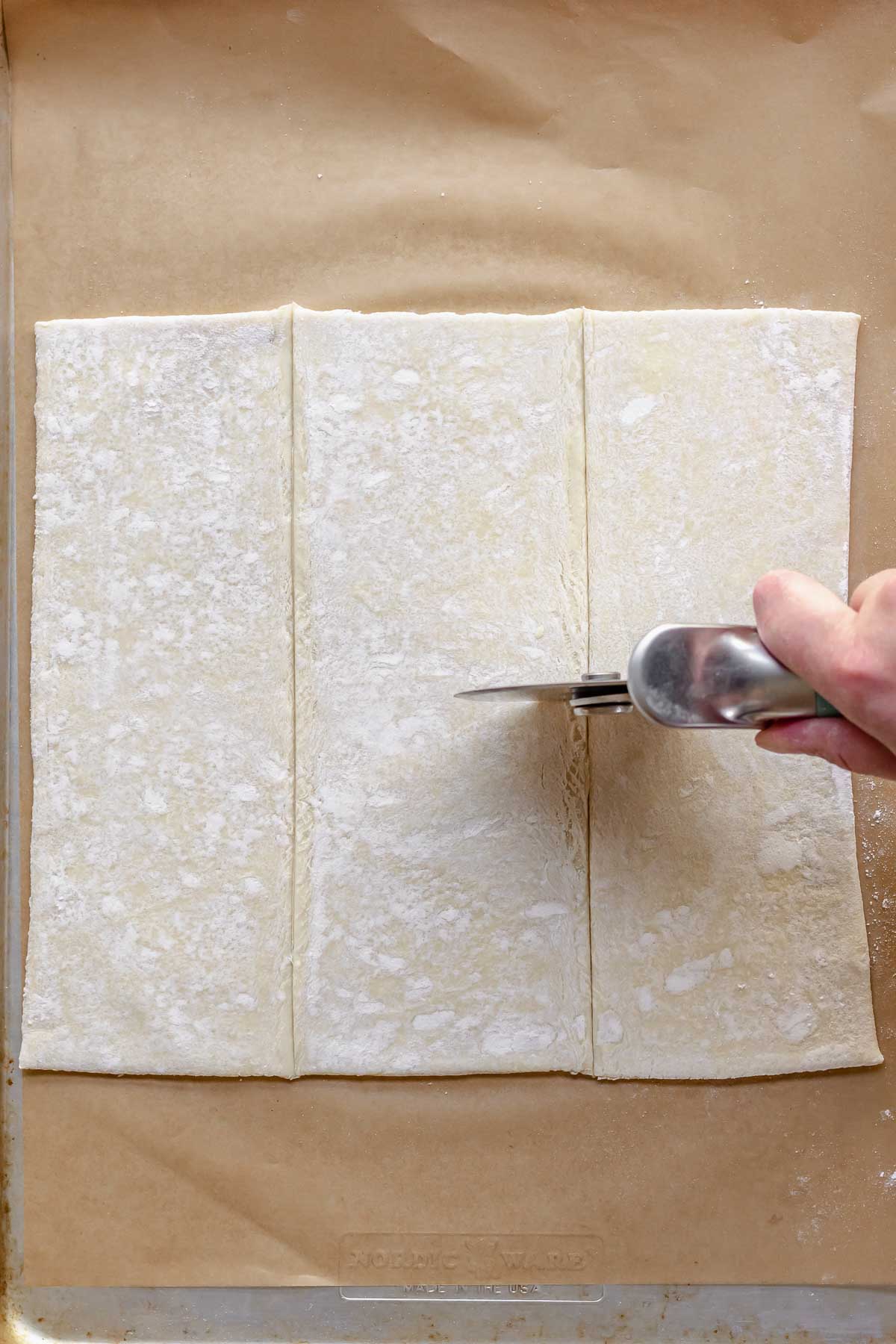 A hand cuts puff pastry into 6 pieces with a pizza cutter.