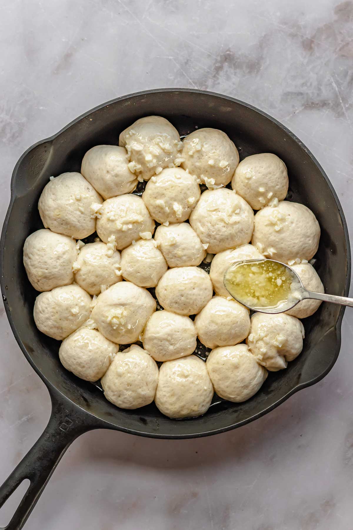 A spoon drizzles melted butter and minced garlic on top of puffy pre-baked bread bites.