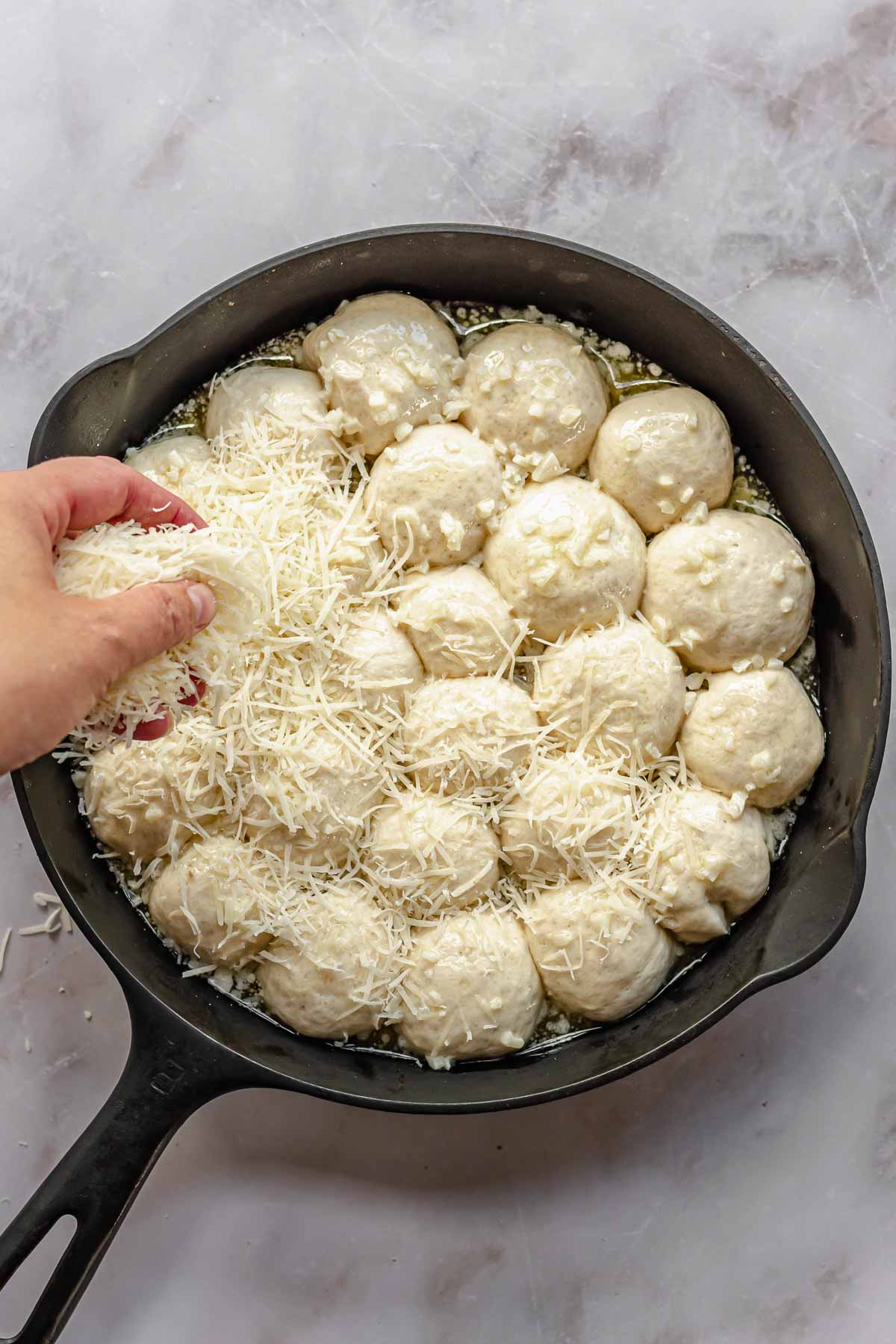 A hand scatters shredded parmesan cheese on top of pre-baked bread bites.