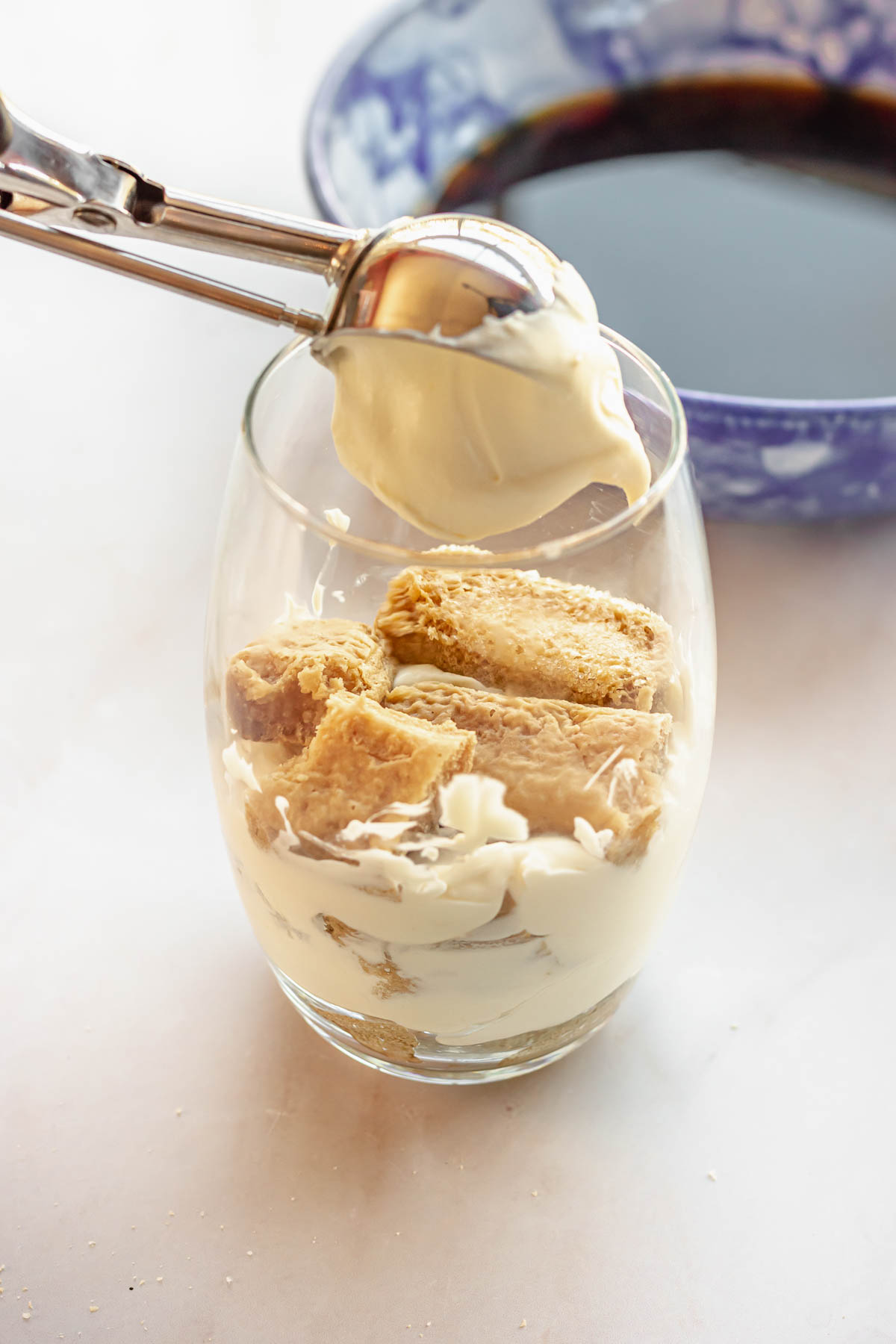 A cookie scoop adds a dollop of cream on top of lady fingers in a cup.