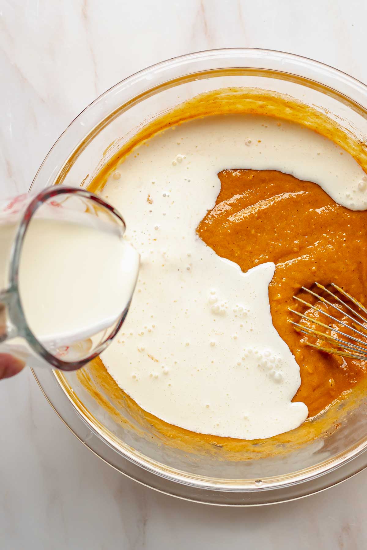 A hand pours heavy cream into sweet potato pie filling.
