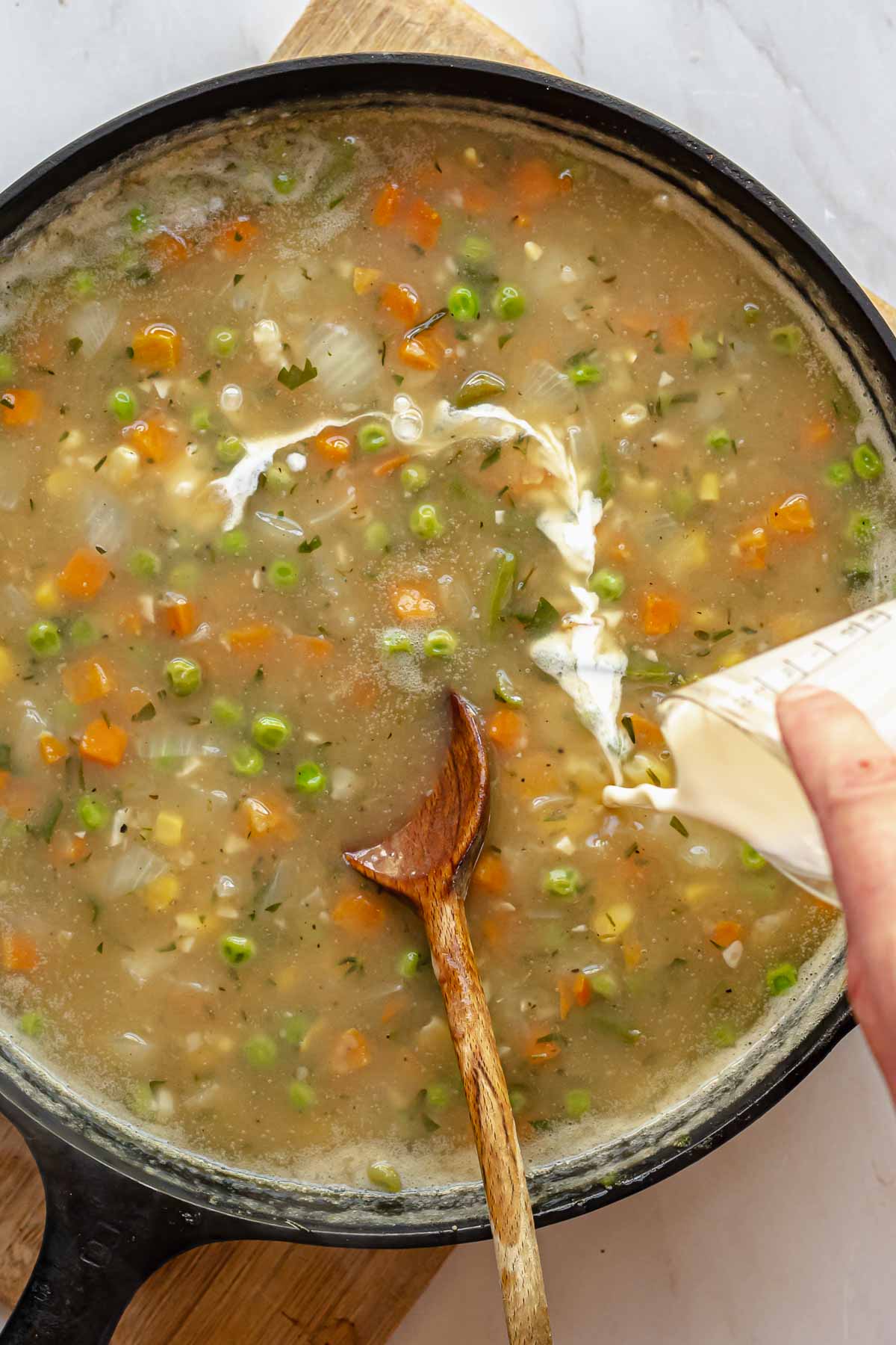 A hand pours heavy cream into vegetables and stock in a pan.