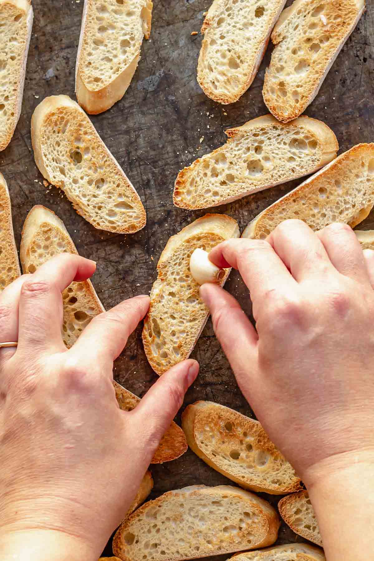 A hand rubs a piece of garlic on toasted baguette slices.