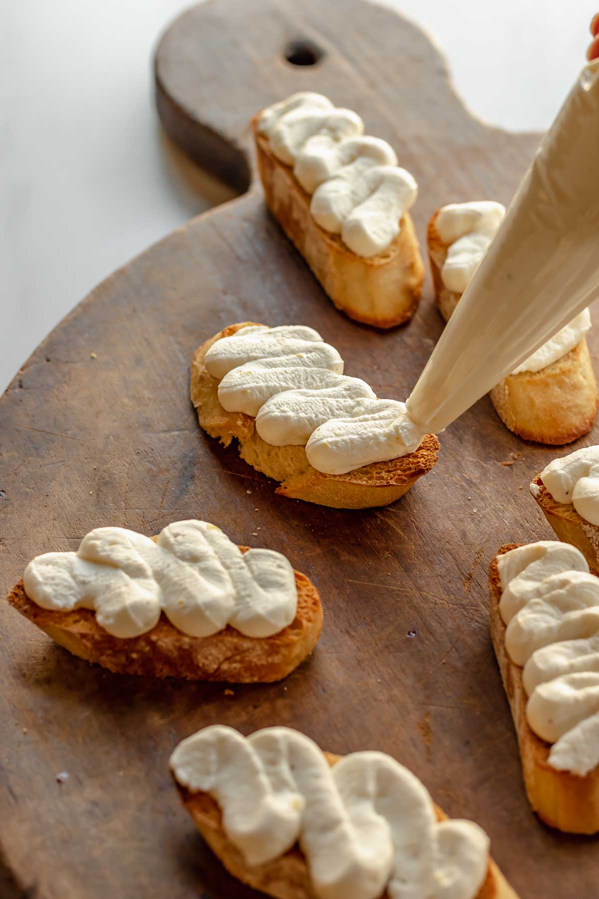 A piping bag pipes squiggles of whipped ricotta onto toasted baguette slices.