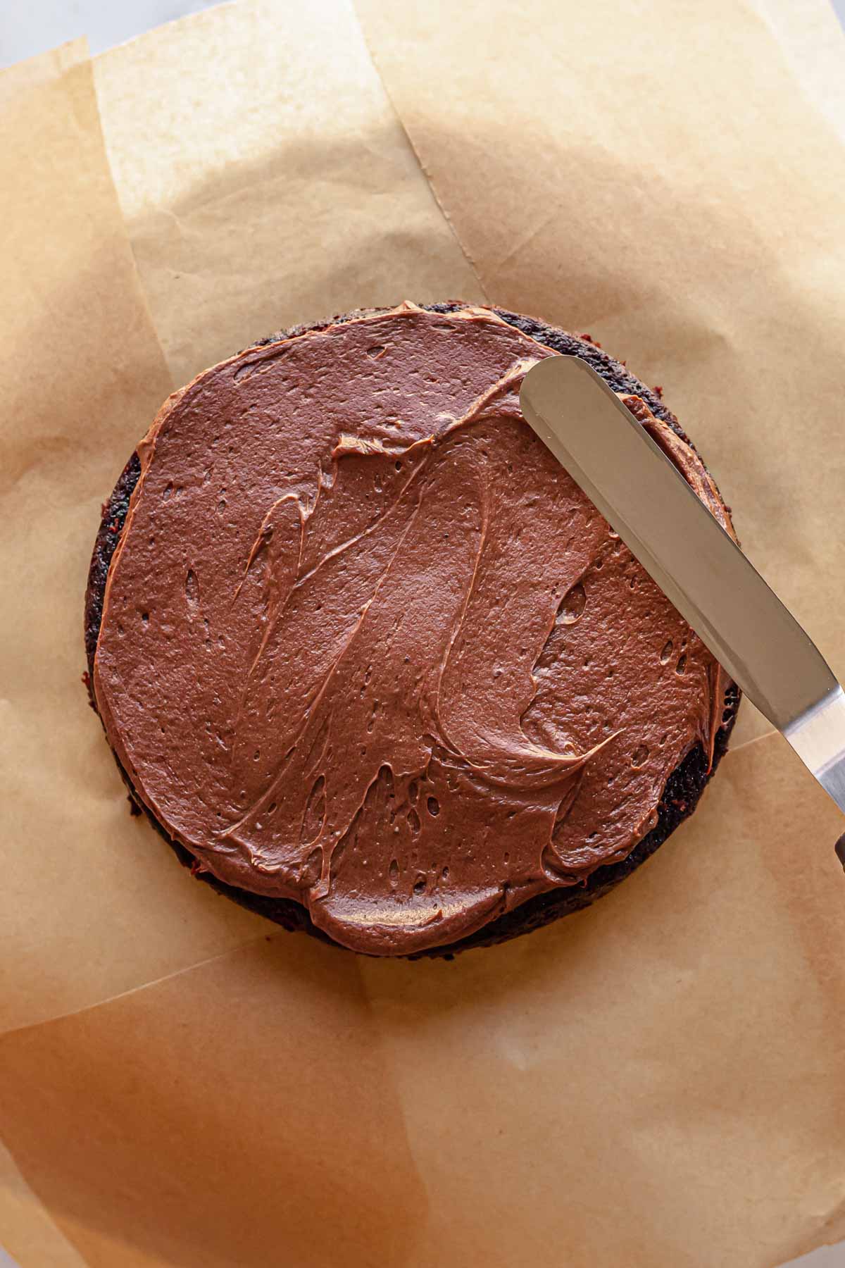 A spatula spreads chocolate cream cheese frosting onto a cake round.