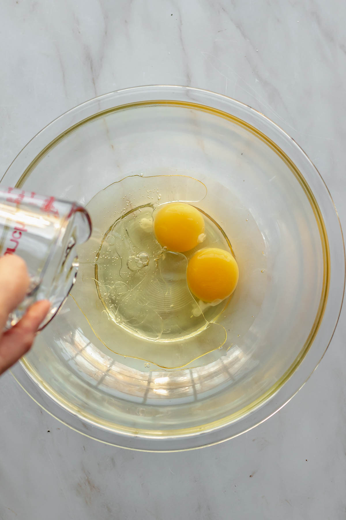 A glass measuring cup pours oil into a bowl with eggs.
