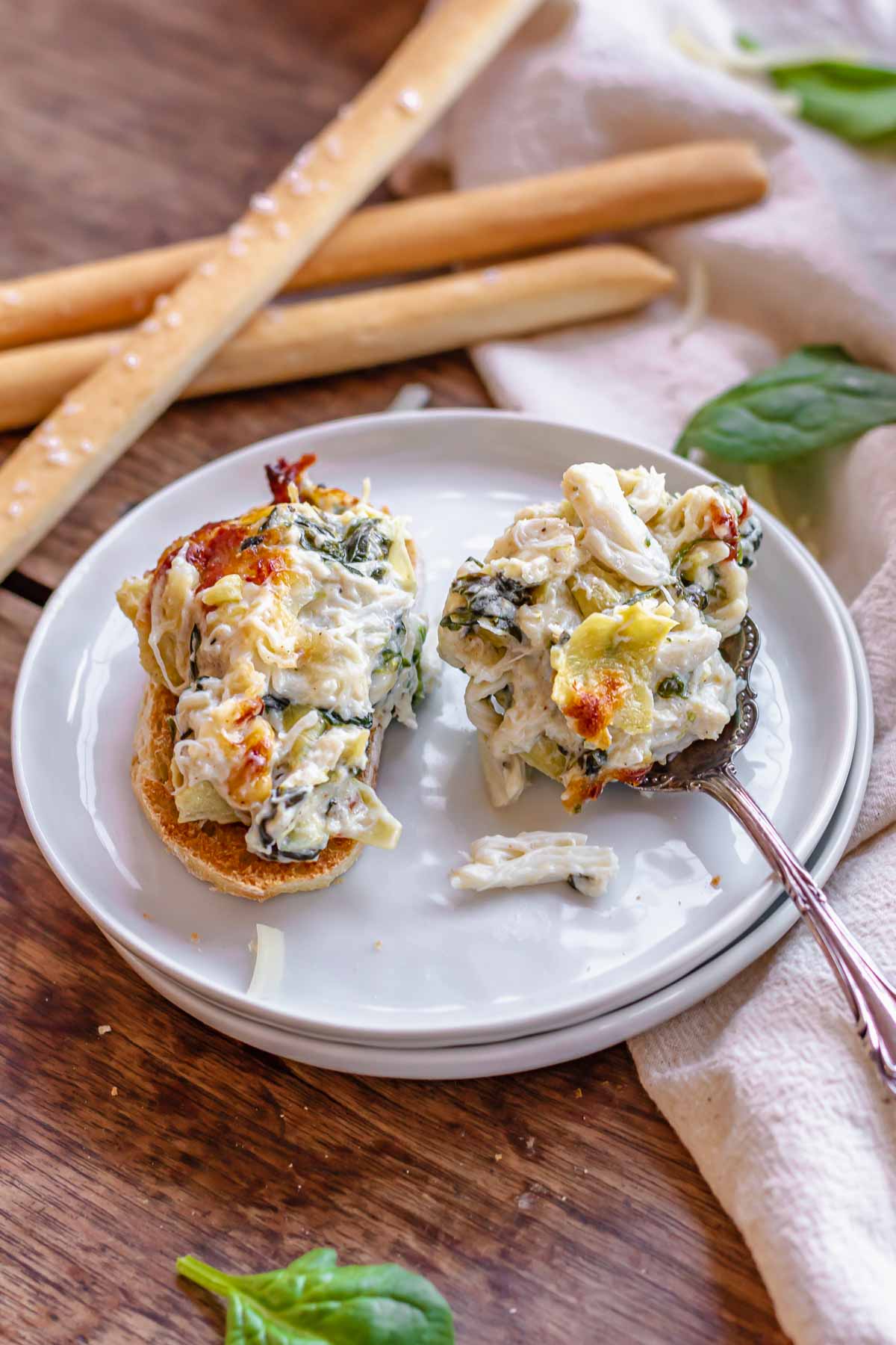 A spoonful of spinach artichoke crab dip on a baguette and some on a spoon resting on a plate.