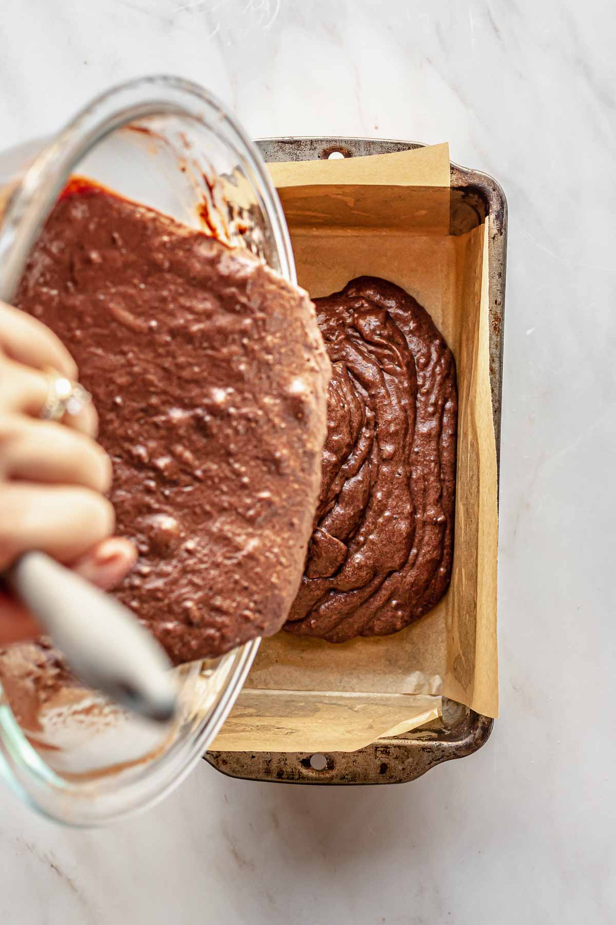 A hand pours chocolate cake batter from a bowl into a loaf pan.