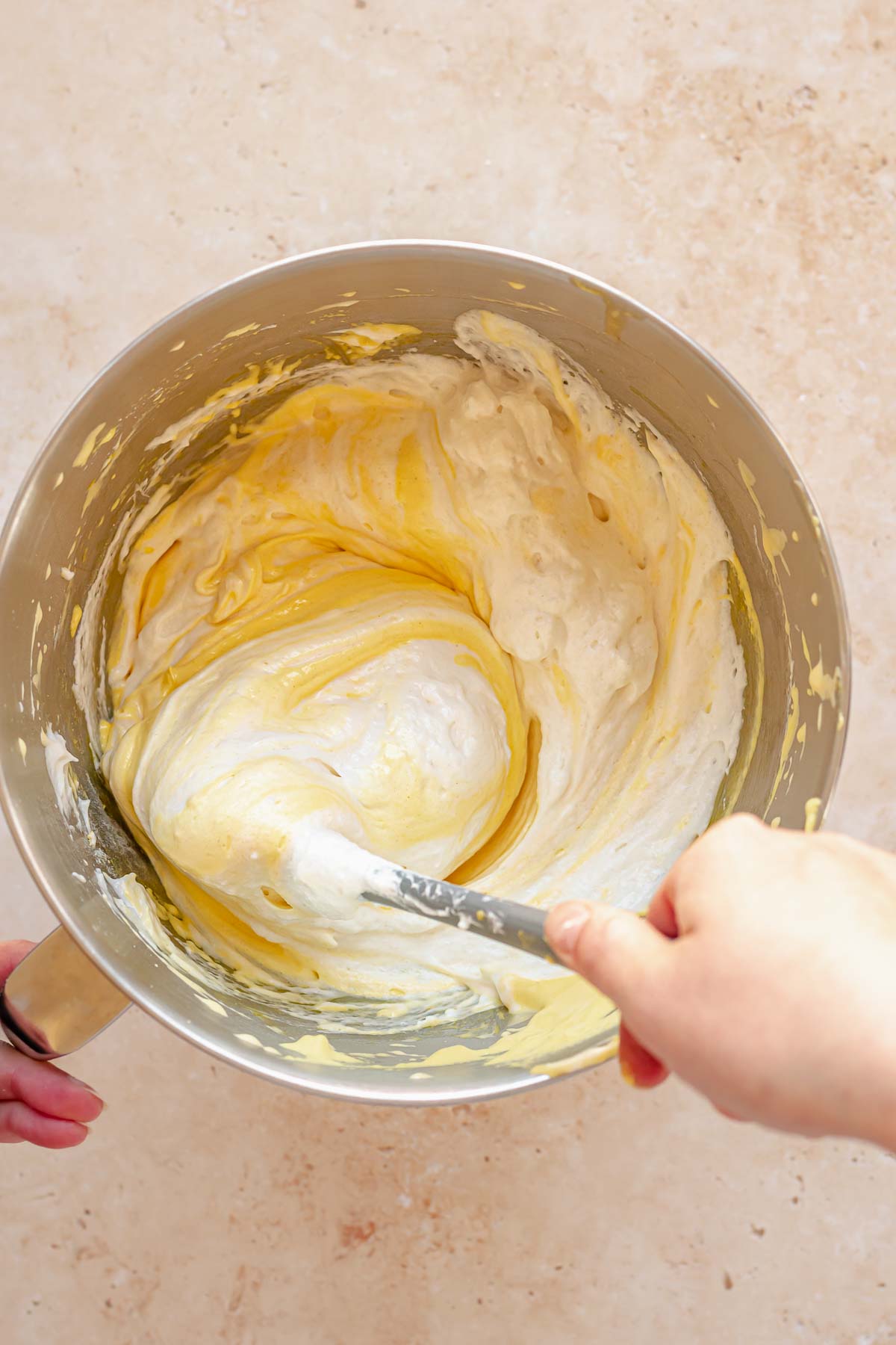 A spatula folds egg whites into egg yolk batter in a bowl.