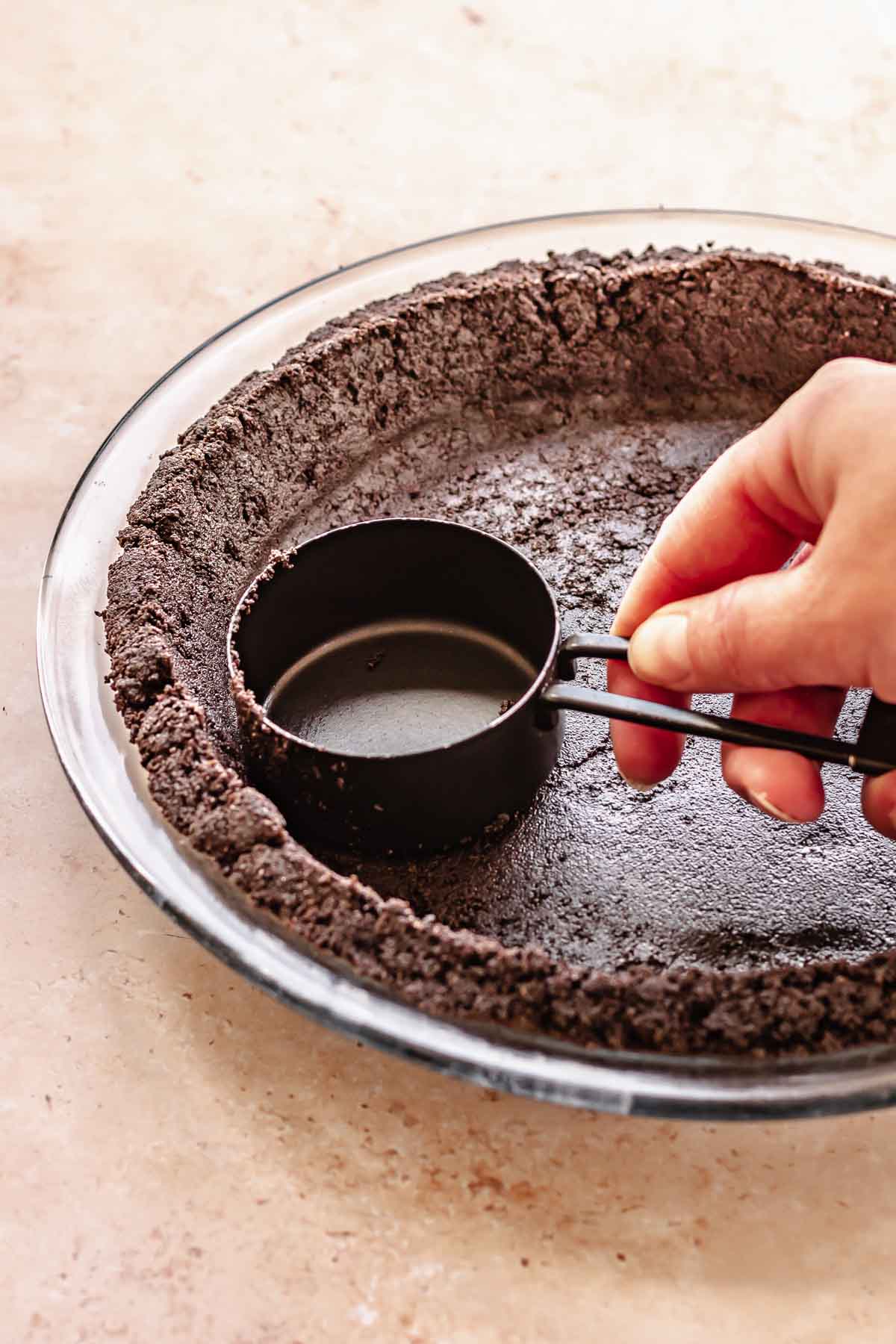 A hand uses a measuring cup to press oreo crust into a pie dish.