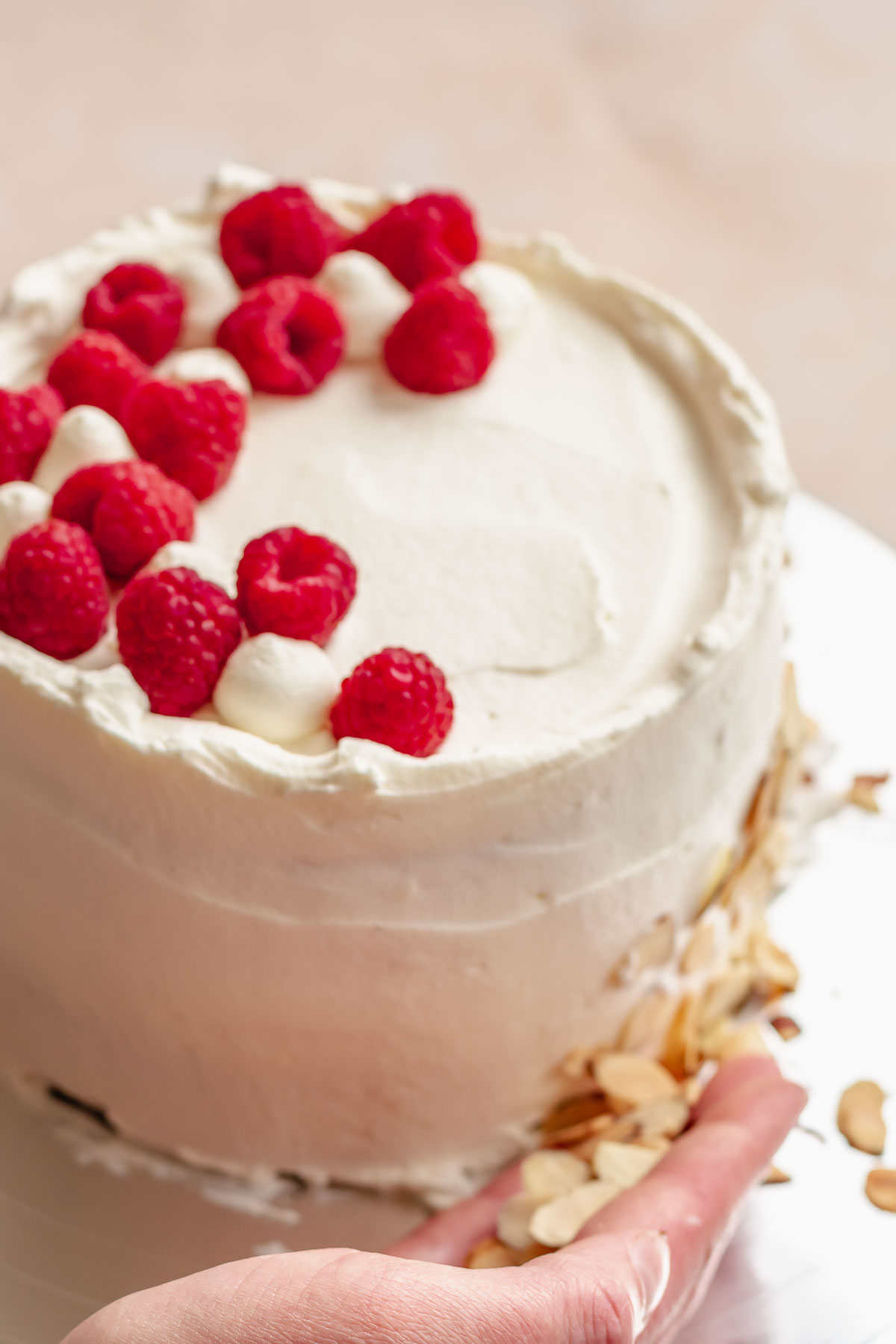A hand presses sliced almonds onto the base of the cake and raspberries are on top.