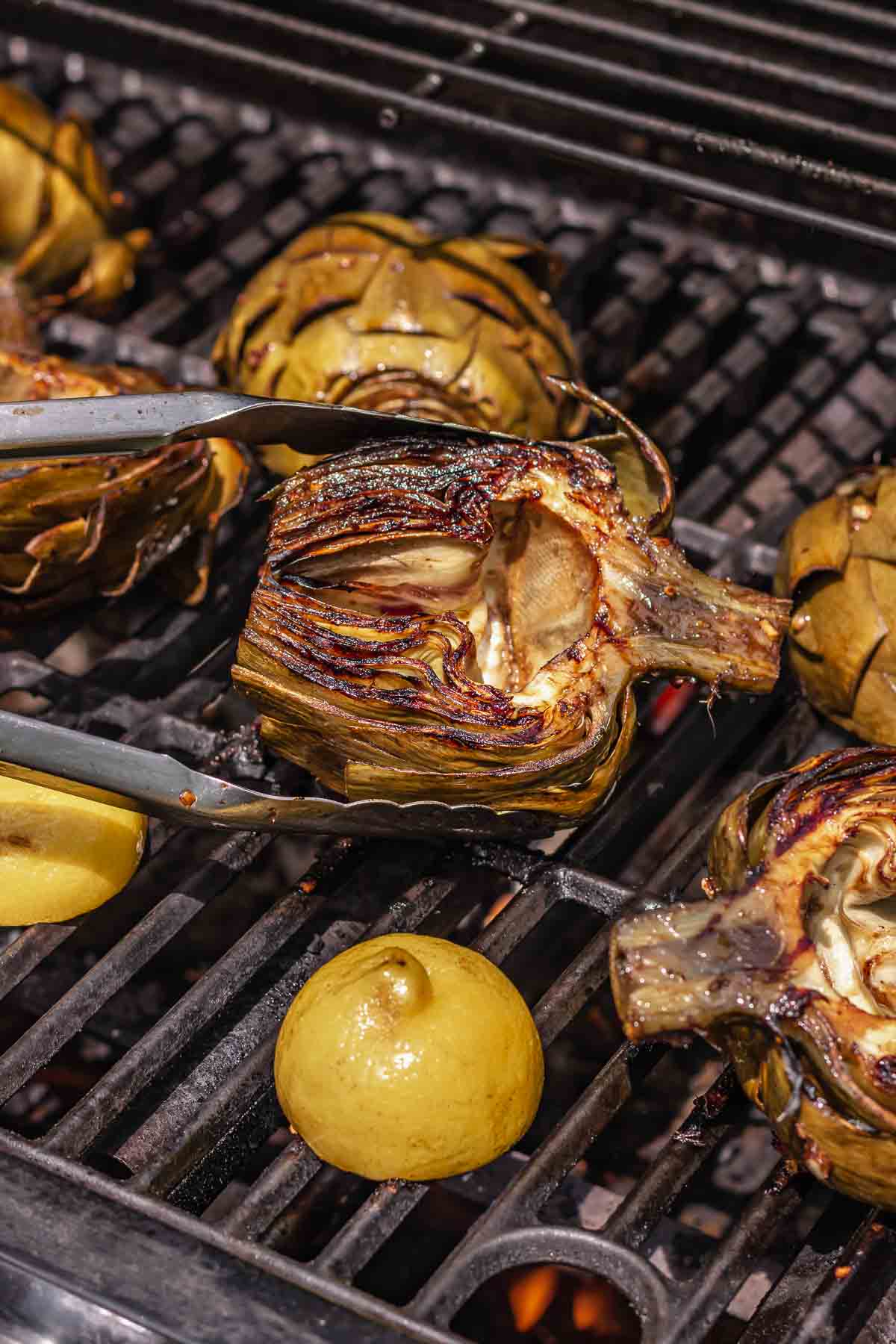 Tongs flip a charbroiled artichoke on the grill.